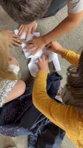 Un groupe d'enfants accroupi au sol, penché sur des morceaux de papier. Ensemble, ils essaient de rassembler les morceaux et déchiffrer le contenu d'une lettre.